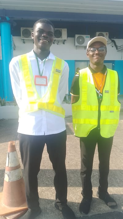 Two young men are standing outside a building in what appears to be an airport environment, likely in Orerokpe, Nigeria, based on the visible "FAAN UGBOKI AIRPORT" logo on their vests. The man on the left is wearing a white collared shirt with a green lanyard and an orange ID card, over which he wears a bright yellow reflective vest. He has a wide, genuine smile. The man on the right is wearing a dark collared shirt with an orange collar, also with a green lanyard and a yellow reflective vest, and a light-colored baseball cap and glasses. He also appears to be smiling. They are standing on a paved surface, with a construction cone to the left of the frame and an air conditioning unit visible on the building behind them. The lighting suggests it is daytime with bright sunlight casting shadows.