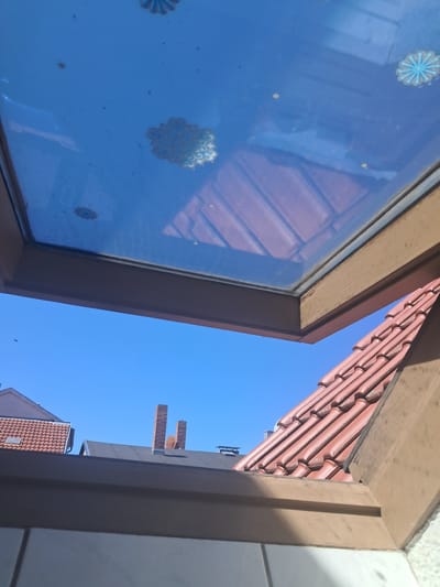 This is a low-angle shot of a window frame looking out onto a clear blue sky. The window has decorative blue and white decals on the glass, resembling snowflakes or mandalas. The wooden frame of the window is visible, as are red roof tiles of a building. In the distance, parts of other buildings and chimneys can be seen against the bright sky. The sun appears to be shining brightly, suggesting it is daytime. There is no text visible in the image. The scene is likely an outdoor setting, possibly a residential area in Parchim, Germany, given the context. The image conveys a sense of a sunny day and an unobstructed view of the sky.