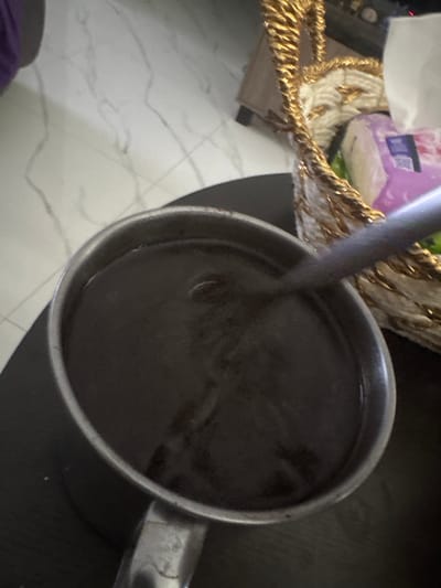 A close-up, overhead shot captures a metal pot filled with a dark liquid, possibly coffee or a similar beverage. The pot has a metallic handle and is placed on a dark, reflective surface. To the right of the pot, a golden-woven basket with white stuffing is visible, partially obscuring some packaging with purple and white hues. In the background, a light-colored, marbled floor is slightly visible. The scene appears to be indoors, with no specific urban or natural features of Sukawati, Indonesia discernible. No people are present in the frame.