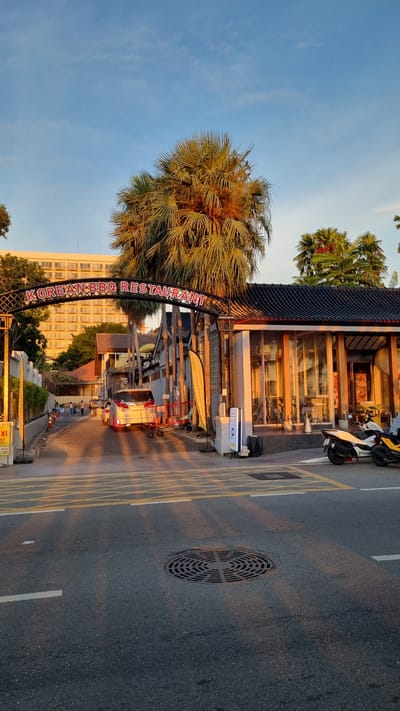 The image shows an outdoor scene, likely a street in Pattaya City, Thailand, captured during sunset, as indicated by the warm, golden light casting long shadows. The sky is a clear, pale blue with a few wispy clouds. Dominating the left side of the frame is a large palm tree with its fronds illuminated by the setting sun.
In the foreground, an archway with decorative metalwork spans across what appears to be a street or pedestrian walkway. Red, illuminated lettering on the arch spells out "KOREAN BBQ RESTAURANT". Behind the arch is a building with a dark, tiled roof, large glass windows, and wooden accents, reflecting the warm light. Several motorcycles are parked in front of the building, one white and black, another yellow.
The street itself is paved and marked with yellow lines. A circular manhole cover is visible on the asphalt. A white van with its brake lights on is driving away from the viewer down the street. Further down the road, other vehicles and a few indistinct figures of people can be seen, suggesting a normal street scene. The overall atmosphere is tranquil and bathed in the soft glow of dusk. The warm lighting and the presence of palm trees contribute to a tropical, evening ambiance.