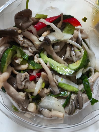 This image shows a close-up of a clear bowl containing a salad. The salad is composed of various ingredients including oyster mushrooms, sliced cucumber, red chili peppers, and what appears to be sliced onions or scallions. The ingredients are mixed together and appear to be dressed in a light liquid. The bowl is partially filled, and the background is blurred, but suggests a simple indoor setting. There are water droplets on the inside of the bowl, indicating it may have been chilled. The overall impression is that of a freshly prepared dish. This image does not provide any information about the location in the city of Asan-si, South Korea.