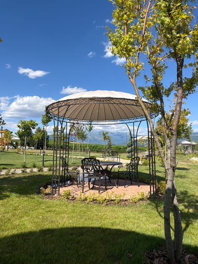 An outdoor scene captures a gazebo with a round, white canopy supported by ornate metal posts in Kocherinovo, Bulgaria. Inside the gazebo, a circular brick patio surrounds a metal table and four chairs. The gazebo is situated on a manicured green lawn with scattered small trees and bushes. In the background, a playground structure is visible to the left, and further back, a vineyard and distant mountains are seen under a bright blue sky with scattered white clouds. A young tree with green leaves stands in the foreground on the right, casting a shadow on the grass.