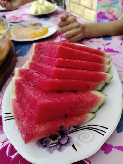 In Galaalti, Azerbaijan, a close-up shot captures slices of watermelon arranged on a white plate with a floral design. The watermelon slices are triangular and stacked, with their bright red flesh and green rinds clearly visible. In the background, out of focus, a person's hands are visible, along with other plates of food, suggesting a meal or gathering. The table is covered with a pink tablecloth featuring a floral pattern.