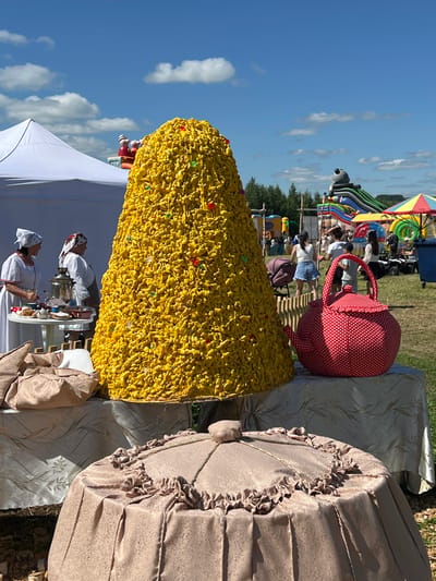 In Kalinino, Russia, on a sunny day with scattered clouds, an outdoor festival is underway. The foreground is dominated by a massive, conical structure, approximately 10 feet tall, covered in thousands of small, yellow, pretzel-like pieces, some adorned with colorful candy. This impressive formation sits on a table draped with a light brown, patterned tablecloth, alongside a large, red and white polka-dot fabric teapot. To the left of the yellow cone, two women in traditional Eastern European attire, including headscarves, stand behind a table laden with a samovar, teacups, and pastries, possibly serving refreshments. In the background, a white tent is visible to the left, and further back, inflatable slides and structures, including one shaped like a panda, suggest a children's entertainment area. Several people are milling about in the background, including families with children. The overall scene depicts a lively community event with a focus on traditional food and entertainment.