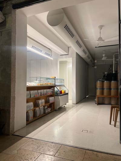 This interior image displays a commercial establishment, likely a coffee shop, located in West Jerusalem, Israel. The foreground features a textured stone floor, which transitions to a smooth, light-colored concrete or tile floor beyond a dark linear threshold.

On the left, a rough stone pillar or wall is visible. The primary space has white walls and a white ceiling from which two large, white cylindrical air ducts with integrated grilles and linear LED lighting are suspended. A third, smaller air duct is partially visible on the far left.

Along the left side, a multi-tiered wooden shelving unit is filled with numerous light-colored, sealed bags, consistent with coffee packaging. Above this unit, a glass-fronted display case exhibits baked goods, situated adjacent to a white refrigerated unit containing various bottled beverages. Further back on the left, a white doorway leads into another brightly illuminated room.

The ceiling above the right section features a long, white cylindrical air duct and two white ceiling fans. A transparent glass partition separates this area from a section on the far right. Within this section, visible through the glass, several cylindrical wooden barrels, some labeled "CAFEDELARTE" and "ESPRESSO," are stacked on a wooden bench. Above them, a large piece of coffee roasting equipment with metal piping is partially visible. A light-colored wooden chair or stool is partially visible in the lower right foreground. No individuals are present in the scene.