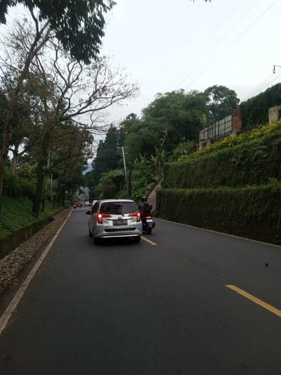A gray minivan with license plate "B 2551 BKN" is traveling on a two-lane road. A motorcycle is to the right of the minivan. The road has a yellow line. A narrow stream bed with leaves is visible to the left of the road. Dense green vegetation covers a hillside on the right of the road. Trees are visible on both sides of the road. Additional vehicles are visible in the distance. The sky is overcast. The location is not Taunggyi, Myanmar.