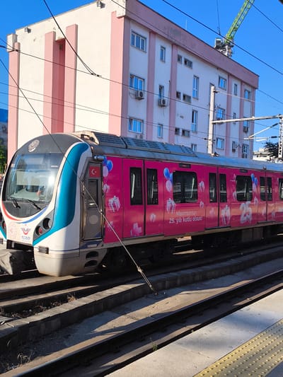 The image shows a train at a station in Pendik, Turkey. The train is decorated with pink and white patterns, including gift boxes and balloons. The text on the side of the train says "Maximum'un 25. yılı kutlu olsun!", which translates to "Happy 25th anniversary of Maximum!". A building with pink and white stripes is visible in the background, along with a construction crane. The sky is blue and clear. The train appears to be stopped at a platform, and the tracks are visible in the foreground.