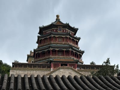 An upward-angled view captures the multi-tiered, octagonal Tower of Buddhist Incense (Fo Xiang Ge) at the Summer Palace (Yihe Yuan), situated in Haidian District, Beijing, China. The pagoda-style structure features dark red pillars, green and blue tiled roofs with distinctive upturned eaves, and intricate detailing. A golden finial adorns the apex, below which a blue plaque with golden characters is visible on the topmost visible level. Several open levels of the tower are discernible, with multiple individuals seen looking out from the balconies or windows on one of the lower levels. In the immediate foreground, the dark, curved tiled roof of another traditional Chinese building is prominent, occupying the bottom portion of the frame. Between this foreground roof and the main tower, a stone wall with a green tiled border and a wooden balustrade is visible, partially obscured by green foliage from trees on both sides. The sky is uniformly overcast and light grey, indicating a cloudy day.