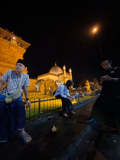 This nighttime image depicts three individuals on a paved walkway in Kauman, Indonesia, with the illuminated Great Mosque of Kauman in the background. On the left, a male subject wearing a black peci, a light-colored patterned short-sleeved shirt, blue sarong-style pants, and grey sandals, sits on a black metal fence. He carries a beige cross-body bag with a green plush attachment and looks slightly forward. In the center, a female subject with dark hair, wearing a white long-sleeved shirt, dark patterned pants, and dark sandals, also sits on the black metal fence, facing left and looking downwards. On the right, a second male subject, dressed in a black long-sleeved shirt with white text and a dark sarong, stands and focuses on a smartphone held in both hands.

The background features the Grand Mosque of Kauman, distinguished by two large central domes, two smaller minarets, and illuminated yellow-golden architectural details. To the left of the mosque, an older, robust stone building with a visible clock face is also illuminated. A bright streetlamp with a starburst effect is present in the upper right of the frame, contributing to the overall artificial lighting. On the dark, paved ground in the foreground, a clear plastic cup, a small clear container holding what appears to be food, and minor debris are visible.