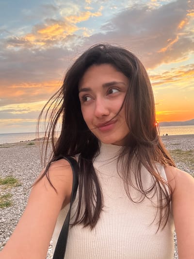 A young woman with long dark hair is pictured at sunset, likely near the beach in Šušanj, Montenegro. She is wearing a light-colored top and has a dark strap, possibly from a bag, over her shoulder. The background shows a beach with pebbles and the sea merging with a sky filled with clouds illuminated by the setting sun. The image is rotated.