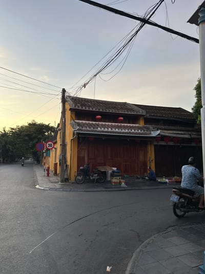 This is a wide shot of a street corner in Hội An Ward, Vietnam. The sky is a pale blue with hints of yellow from the setting sun. The street is paved and asphalted, with a white line marking the center of the road in some areas. There are several buildings lining the street, most notably a yellow building with a red-tiled roof and traditional wooden doors. Red lanterns are hung under the eaves of the building.

In front of the yellow building, there are several objects, including a motorcycle, a wooden table, and various goods that appear to be for sale. Two people are present, one sitting on the ground near the table and the other, a person wearing a blue patterned shirt and shorts, riding a motorcycle away from the camera. A red fire hydrant is visible on the sidewalk near a street sign that indicates a speed limit of 30.

Trees with dense green foliage line the street on the left side of the image. Power lines are strung overhead, crisscrossing the sky. The overall impression is of a quiet street in a historic Vietnamese town as dusk approaches.