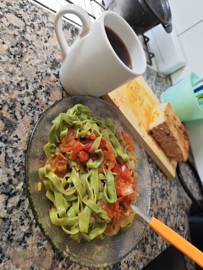 The image presents a meal setting on a countertop with a speckled granite-like surface. A clear glass bowl contains green-colored pasta mixed with diced tomatoes and onions. Beside the bowl is a white mug filled with a dark liquid, presumably coffee, and a wooden cutting board with a slice of brown loaf or cake on top. An orange-handled utensil is partially submerged in the pasta. In the background, a small appliance is visible. The location is unspecified but inferred to be La Plata, Argentina. There is no explicit evidence of people present in the image.