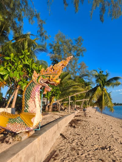 A vibrant, brightly colored statue of a mythical Naga serpent, with an ornate headdress and scales, stands on a sandy beach in Baan Lipa Noi, Thailand. The serpent is positioned in the foreground, partially obscuring a lush green tree and palm trees. Behind the statue, a concrete wall runs along the edge of the beach, separating it from a path lined with more palm trees. In the distance, several swings hang from wooden frames, and a lone person walks along the shoreline. The ocean, a calm blue, stretches to the horizon under a clear, bright blue sky. The sand on the beach is light brown and scattered with small debris. The overall scene depicts a tropical beach setting with a touch of cultural artistry.
