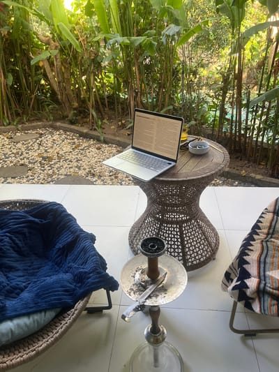 The image shows a laptop on a wooden table in an outdoor setting. The table is next to a stone garden. A hookah sits on the ground next to a brown woven chair with a blue blanket. The scene is in Kerobokan, Indonesia.