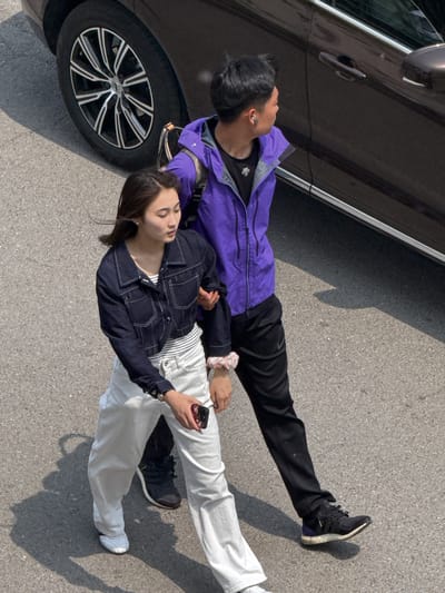 An aerial view shows a man and a woman lying on the asphalt next to a parked car. The man is wearing a purple jacket and black pants, with black sneakers. He is lying on his back, near the car's front driver-side tire. The woman is lying on top of the man, wearing a dark blue jacket, a striped shirt, and white pants with white sneakers. She is holding a cellphone in her hand. The location appears to be Zhongtun, China.