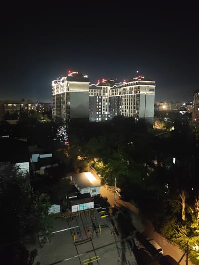 An aerial, nighttime, long-shot shows a modern, illuminated residential complex in Bishkek, Kyrgyzstan. The complex has at least two tall, multi-story buildings with numerous windows lit from within. The buildings are crowned with decorative lighting and red rooftop markers. A large sign on one of the buildings reads "ELITE HOUSE". Surrounding the complex are other, less illuminated buildings, trees, and a playground area with swings and slides. A street with parked cars and a few moving vehicles is visible. The overall scene depicts a well-lit urban residential area at night.
