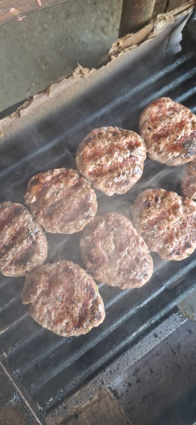 This image captures a close-up, top-down view of several grilled patties, likely made of ground meat, arranged on a black metal grill. The patties are browned with distinct grill marks, indicating they are being cooked. Wisps of steam rise from the grill, suggesting the patties are hot. The grill itself appears to be outdoors, with some weathered wooden elements visible at the top and a concrete or dirt surface below. The overall impression is of a casual outdoor cooking environment. The image does not provide any specific visual cues to pinpoint its location within Lyaskovets, Bulgaria.