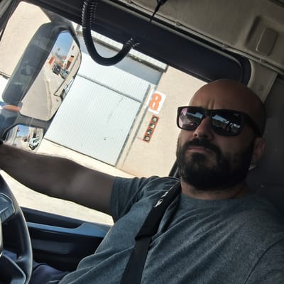A man with a beard and sunglasses is in the driver's seat of a truck. He is wearing a gray t-shirt and a seatbelt. The truck's side mirror reflects a street in Nàquera, Spain, and a building numbered "8" is visible through the windshield.