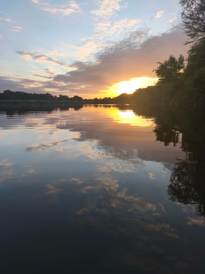 The image depicts a serene sunset over a body of water, likely a river, with the city of Saratovskii, Russia, in the background. The sky is a canvas of soft blues and oranges, with wispy clouds reflecting on the calm water. Lush green trees line the banks, their silhouettes stark against the vibrant sky. The overall mood is peaceful and tranquil, capturing the beauty of nature at dusk.