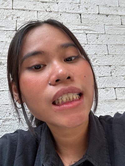 A close-up, eye-level, outdoor selfie shows a young person in South Jakarta, Indonesia, against a white brick wall. The subject, with dark hair, is wearing a black collared shirt and has makeup on, including blush and eyeliner. Their mouth is slightly open, revealing teeth that appear discolored. The lighting suggests it is daytime.