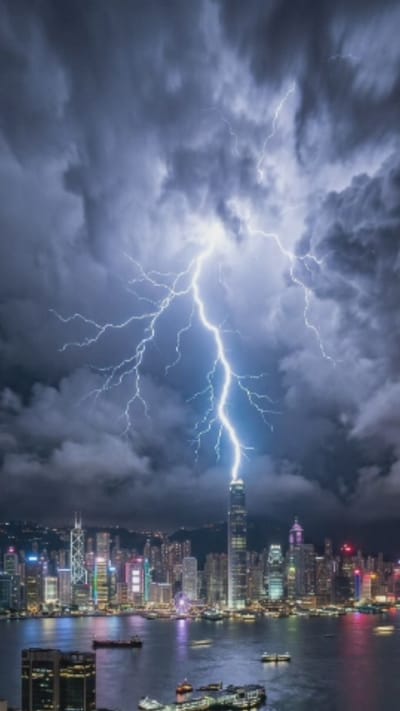 This media file depicts a dramatic nighttime thunderstorm over Hong Kong's iconic skyline. The primary subject is the vibrant city illuminated by a powerful lightning strike that appears to hit a very tall skyscraper, possibly the IFC Two building, situated prominently in the center of the frame. The lightning, a brilliant white and blue jagged line, forks out as it descends from dark, swirling storm clouds that fill the upper portion of the image.

Below the lightning and clouds, the city sprawls across the horizon, a dense collection of illuminated skyscrapers. The buildings display a variety of colors from their lighting, including white, blue, green, and pink, creating a festive yet intense atmosphere. Notable structures visible include other prominent skyscrapers, a Ferris wheel, and the general urban landscape. The scene is set on the water, likely Victoria Harbour, with several ships and boats, including cargo ships and ferries, visible on its dark surface. The water reflects the city lights, adding to the dynamic visual.

The time of day is clearly night, as evidenced by the artificial lighting of the city and the dark, stormy sky. The weather is extreme, characterized by the powerful thunderstorm and lightning. The location is unmistakably Hong Kong, identifiable by its characteristic skyline and the geographical layout of its harbor. The overall mood is one of awe and power, with the raw energy of nature juxtaposed against the man-made marvel of the city. There is no visible text in the image.