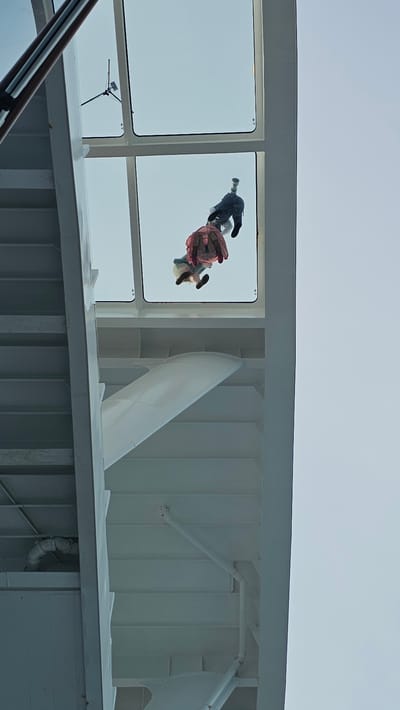 The image is taken from a low angle, looking up through a window or glass panel of a ship or building, seemingly in the city of Marseille, France. Two people are visible through the window, appearing distorted due to the viewing angle. One person is wearing dark clothing and the other is wearing a pink dress, both figures captured from below. The surrounding structure is white, with metal beams and a life raft on the ship. The sky is visible through the window.