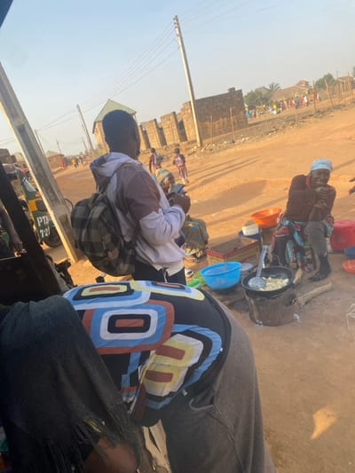 The image depicts an outdoor scene in Udu, Nigeria, characterized by a bustling roadside environment under clear daylight. In the immediate foreground, the back of an individual wearing a dark shirt with large, colorful geometric patterns and grey trousers is visible, partially obscuring the lower left of the frame.
The midground features a man standing with his back mostly to the camera, wearing a white and brown hooded sweatshirt and a plaid backpack. He holds a smartphone in his right hand. To his right, an older woman is seated on a low stool, wearing a blue headwrap, a brown top, and a brightly patterned skirt. She is smiling and looking towards the viewer's general direction. Next to her, a cooking station is active: a dark pot containing items (possibly plantains or yams) frying in oil sits on a makeshift stove, likely fueled by wood or charcoal, with visible flames. A metal slotted spoon rests in the pot. Nearby are colorful plastic bowls, including an orange and a light blue one, along with a red lidded container.
The ground across the scene is unpaved, dry reddish-brown dirt. In the background, a dirt road stretches into the distance, with several individuals, including children, walking on it. Partially constructed buildings made of concrete blocks, some with exposed rebar, line the right side of the road, separated by a fence. Utility poles with overhead power lines are visible throughout the scene. On the far left, part of a green and yellow tricycle (Keke Napep) with "PTA 721" text is discernible. The sky is bright and clear, casting distinct shadows.