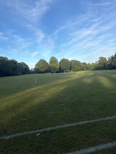This image displays an outdoor grass sports field, likely a football pitch, situated in Maidstone, United Kingdom. The field is marked with white lines, including a prominent line across the foreground and another perpendicular line further back. Three goal frames are visible: a full-sized white goal with a net positioned on the right mid-ground, and two smaller, white or light-colored goal frames without nets, located further left and center-left on the field. The periphery of the field is lined with mature deciduous trees, exhibiting dense green foliage. Above, the sky is clear blue with sparse, wispy white clouds. Long shadows from an unseen source on the left extend across the grass, indicating a low sun angle. No individuals, animals, or active events are present within the frame. Scattered dry leaves are visible on the grass in the foreground.