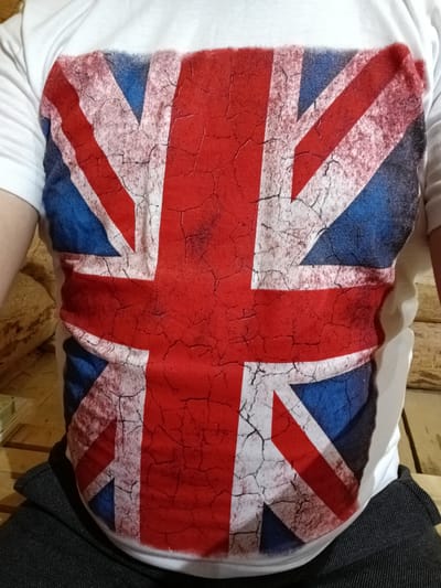 The image shows a close-up of a person wearing a white t-shirt with a distressed print of the Union Jack flag. The flag design features red, white, and blue colors with a crackled, vintage effect. The person is wearing dark gray trousers. The background appears to be a wooden surface, possibly a table or bench, with some soft, textured material to the left, which could be upholstery. The image is tightly cropped and does not provide any discernible information about the location in the city of Молочное, Russia.