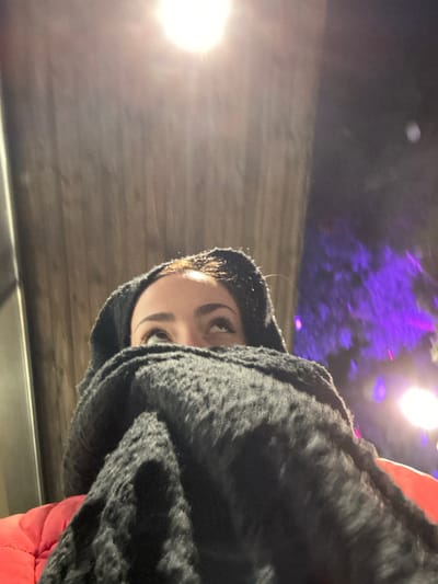 A person is closely framed, their face partially obscured by a black, furry hood or shawl. Only one eye and part of their forehead are visible, looking upward. The lighting is unusual, with a bright light source on the left side casting strong highlights. The background features a wooden or textured surface, potentially a wall or ceiling, with additional, blurred lights and hints of purple foliage above. A glimpse of red fabric suggests an article of clothing.