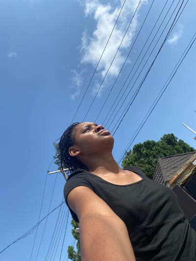 A low-angle, daytime shot shows a person with brown skin and braided hair, wearing a black shirt. The person is looking upward. The background features a blue sky with light clouds, power lines, a green tree, and part of a building with a roof. The location is Iranla, Nigeria.