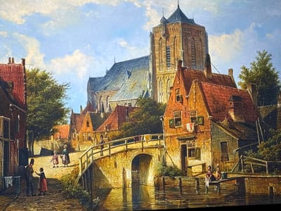 This painting depicts a detailed Northern European canal-side town scene, characterized by a Gothic-style architectural aesthetic not consistent with the city of Reus, Spain. The prominent structure in the background is a large, multi-tiered brick church or cathedral featuring a tall, spired tower and a grey slate roof, bathed in bright sunlight.

In the foreground, a cobblestone street leads towards a stone arch bridge spanning a canal. On the left, two figures stand near a building: a man in a dark uniform and hat faces a child in a red dress who gestures towards him. White laundry hangs on a line near this building.

On the street beyond the bridge, three figures, two women and a child, walk towards the bridge. One woman wears a green apron and carries a basket. Another lone figure in dark clothing walks away from the viewer on the bridge itself.

To the right, several multi-story brick houses with characteristic red tiled roofs and green-shuttered windows line the canal. Laundry, including white and colored garments, hangs from lines outside the windows of the houses immediately adjacent to the bridge. At the water's edge, on a wooden pier, two women are engaged in activity: one is seated, seemingly washing clothes with a white cloth, while another stands nearby with a bucket. The tranquil canal water reflects the surrounding buildings and sky. Green trees are interspersed throughout the urban landscape. The scene is set under a bright blue sky with scattered white clouds.