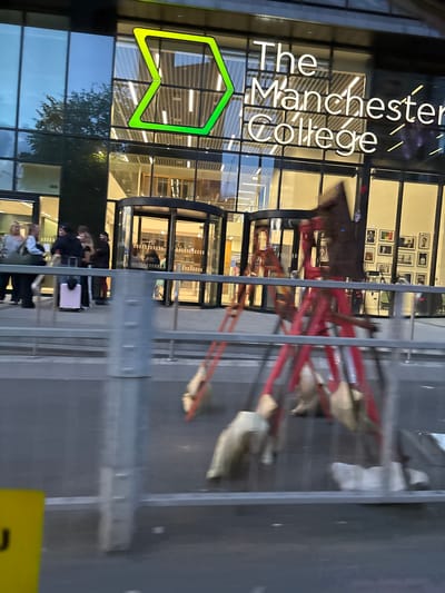 The image captures the exterior of The Manchester College in Manchester, United Kingdom. The college's signage is prominently displayed, featuring the name "The Manchester College" in white lettering with a green logo. The building's façade is predominantly glass, reflecting the surrounding environment. A group of people is visible near the entrance, some with luggage, indicating potential students or visitors. A metallic barrier is in the foreground.