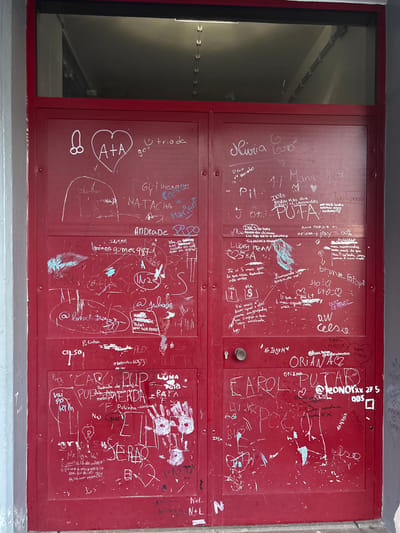 The image depicts a red double door covered in graffiti. The door is likely located in Câmara de Lobos, Portugal. Various names, phrases, and drawings are scrawled on the surface. A partially visible window is adjacent to the doors. The environment suggests an urban setting with signs of wear and tear.