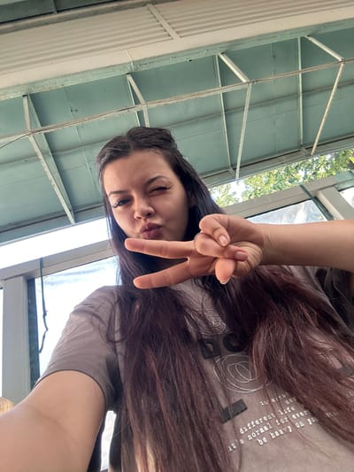 A young woman with long, dark brown hair is taking a selfie from a low angle. She is wearing a gray t-shirt. Her left hand is raised, making a peace sign with her index and middle fingers. Her right hand is not fully visible. She is puckering her lips and winking. The background appears to be an outdoor covered area, possibly a patio or balcony, with a slatted ceiling and visible structural beams. Sunlight filters through foliage in the background. The overall impression is a casual, personal photograph. Without further context, the specific location within Sliven, Bulgaria, cannot be determined from the image alone.