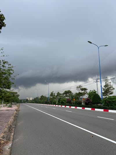 The image depicts a wide, paved road in Phan Thiết, Vietnam, with a single white line marking the center lane and a white line with alternating red and white segments bordering the right side. The road stretches into the distance, lined with trees and shrubs on both sides. On the left, there is a paved pedestrian walkway with a concrete curb. On the right, the road is bordered by a low white wall topped with red and white segments, followed by a row of lush green bushes, some with pink flowers, and trees. Several streetlights with dual lamps on blue poles are visible along the road. In the background, there are more trees and some buildings. The sky is overcast with dark, heavy storm clouds dominating the upper portion of the image. A single vehicle is visible in the far distance on the road.