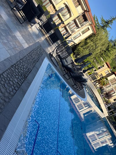 The image depicts a poolside scene at a resort or hotel in Sandanski, Bulgaria. In the foreground, a swimming pool with blue mosaic tiles is visible, with a reflection of a building on its surface. The pool is bordered by a white tiled edge and a section of decorative cobblestones. A row of black lounge chairs and umbrellas are set up on a tiled patio area to the left of the pool. Beyond the patio, a multi-story building with yellow and beige facades, red-tiled roofs, and numerous windows is visible. Lush green trees and foliage surround the building, with a clear blue sky overhead. The overall scene suggests a tranquil, sunny day at a vacation destination. There are no people visible in the image.