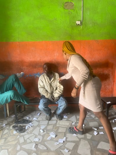The scene is an indoor space in Sabon Gari East, Nigeria. The room features a distressed two-toned wall; the upper half is painted lime green, and the lower half is orange. Both sections display significant wear, dirt, and areas of peeling paint, with notable blue discoloration along the lower orange section. An electrical switch or outlet is mounted on the upper right portion of the green wall. The floor is tiled with irregular, light-colored mosaic patterns and is covered with numerous scattered small, white, rectangular pieces of paper or wrappers.

Three individuals are present. On the left, the lower legs of a person, clad in teal green trousers, are visible, seated on a metal bench with a black frame and light-colored top. Centered in the frame, an older adult male, with dark skin and a bald head, is seated on the same bench. He wears a light-colored, possibly beige, long-sleeved jacket and blue trousers, with dark sandals. His mouth is open, indicating speech or a reactive expression, as he looks towards the right. To the right of the seated male, an adult female with long, bright yellow braided hair stands, leaning forward and facing him. She wears a light beige long-sleeved dress and black platform sandals with red sole accents. Her right arm is extended, and her right hand or finger is directed towards or touching the male's upper left arm or shoulder area. A dark beaded bracelet is visible on her right wrist. Two black slip-on sandals are on the floor near the left side of the metal bench. A small, dark table or stool is partially visible on the far right.