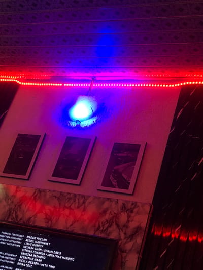 The image shows a wall with three framed pictures and an LED light strip. The light strip emits red light at the top and blue light below, creating a colorful ambiance. The framed pictures appear to be abstract art or possibly photographs. In the lower part of the image, a dark screen displays text, which seems to be a list of names and job titles, possibly credits from a film or production. The wall has a textured surface, with some parts appearing to be marble-patterned. The overall impression is that of an entertainment venue or a place decorated with a modern, artistic theme, possibly in New Stadium, Nigeria. There are no people visible in the image.