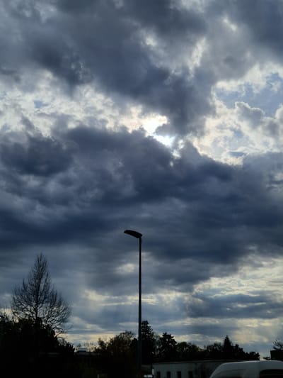 The image depicts a view of a cloudy sky with a street lamp in the foreground. The location is Rheinfelden, Switzerland, based on contextual clues. The sky is filled with dark, heavy clouds with glimpses of sunlight breaking through. A street lamp extends horizontally across the frame, with its curved head facing downward. Bare trees and the edge of a building are visible in the background. The scene appears to be taken during the daytime.