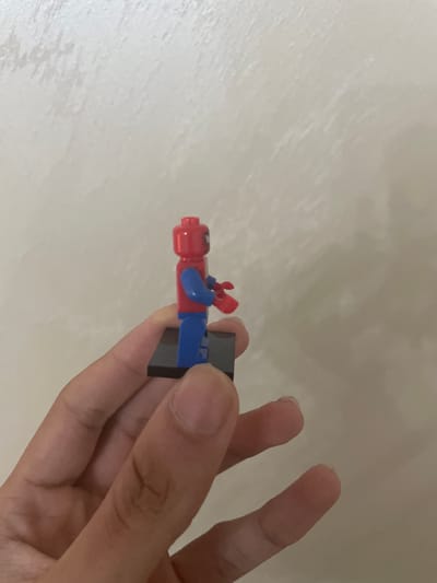 The scene depicts a human hand holding a Lego minifigure against a plain, textured wall, occurring in the city of Ihtiman, Bulgaria. The hand, with light to medium skin tone, occupies the lower portion of the frame, with visible fingers and knuckles. The Lego minifigure is predominantly red and blue, featuring a red head with indistinct facial markings, a red torso with blue arms, and blue legs. The minifigure's left arm is bent, and its hand holds a small, red, indistinct accessory. It stands on a rectangular black Lego display base (2x3 studs). The background is a uniformly lit, light beige or off-white wall with subtle, irregular surface texture.