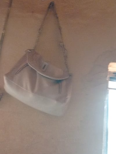 The image displays an indoor scene within Nsukka, Nigeria. A two-toned shoulder bag is suspended by a thin metallic chain strap against a light brown wall. The bag's upper section is light brown, while its lower portion is off-white, and it features a front flap closure with a small metallic accent. A bright, vertical light source, consistent with a window or doorway, is visible along the right edge of the frame. The overall image exhibits a grainy texture and slight blur. No individuals or other distinct objects are present.
