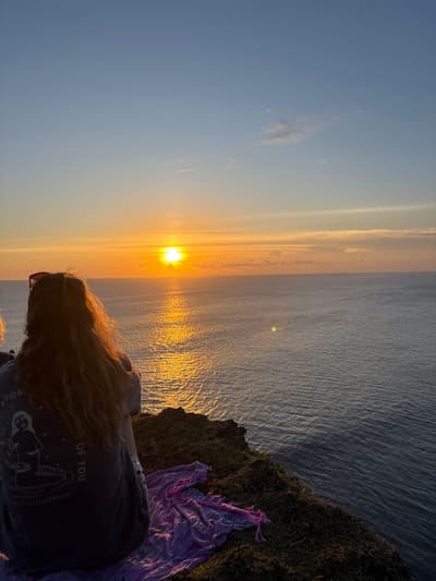 A woman with long blonde hair is standing on a cliff overlooking the ocean at sunset in Pecatu, Indonesia. The sun is setting over the water, creating a golden reflection on the surface. The cliff is covered in grass and there is a purple cloth draped over the edge. The woman is wearing a dark t-shirt with a graphic on it.