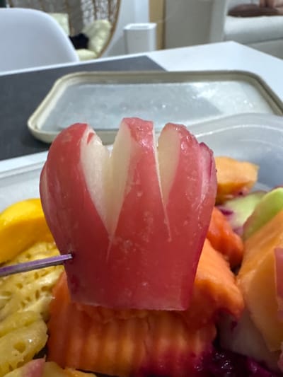 In a shallow plastic container filled with a variety of colorful cut fruits, a single rose apple is prominently displayed. The rose apple, a vibrant pinkish-red, has been artfully carved to resemble a blooming flower, with four distinct petals. It is pierced by a small, thin purple skewer, hinting at its presentation as a fruit skewer. Surrounding the rose apple are chunks of other fruits, including bright yellow mango, orange cantaloupe, and what appears to be pineapple and possibly dragon fruit due to the deep purple accents. The fruits are arranged closely together within the clear container. The background suggests an indoor setting, possibly a dining area, with a blurred view of furniture and a tray. The overall scene captures a close-up, detailed view of a fruit arrangement, highlighting the meticulous preparation of one of the fruits. This image does not provide any specific information about the location in Kathu, Thailand.