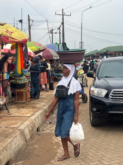 A market scene unfolds in Egan, Nigeria. A woman balances a large brown bowl on her head while walking along a brick-paved street. She wears a white headscarf, a white top, a denim skirt, sandals, and has a black bag slung across her body. She holds a white plastic bag in her right hand.
To her left, market stalls are covered with colorful umbrellas and display various goods. Other people are browsing or selling at the stalls. A mannequin head with long black hair is visible among the goods.
To her right, a black SUV is parked at the edge of the road. The street appears crowded. Power lines crisscross overhead against a cloudy sky. A concrete curb separates the street from a lower, dirtier area.