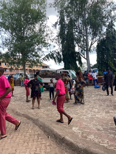 The image depicts an outdoor scene in what appears to be a public area within the city of Nnewi, Nigeria, likely a school or community gathering space, judging by the presence of young people in uniform attire. The foreground is dominated by a reddish-brown patterned paved walkway. Several individuals are present, most notably two young males in the foreground wearing bright pink shirts and dark red trousers. They are walking or standing, with their backs partially to the camera.
In the midground, a diverse group of people are congregated. Several women are wearing colorful, patterned dresses and headwraps. One woman in a black dress with red polka dots stands facing the camera, while others are engaged in conversation or observing their surroundings. A young boy in a gray t-shirt and black trousers stands near a white truck.
The background features a light-colored building with multiple windows and a multi-story structure. Several vehicles are parked in the vicinity, including a white truck with what appears to be wood or lumber in its bed, and other cars. Tall trees with dense green foliage line the area, and the sky is overcast with some lighter patches. The overall impression is one of a lively gathering or event taking place outdoors.