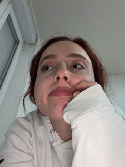 A young woman with auburn hair and light skin strikes a selfie pose. She's wearing a white hoodie, her hand playfully squishing her cheek. Her eyebrows are thick and well-defined, and her eyes are wide and expressive. Her lips are full, and there appears to be a subtle shine on her face. The picture is taken from a slightly low angle, emphasizing the woman's face, and a window is visible in the background.