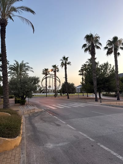 The image depicts a street scene in Benidorm, Spain, at what appears to be dusk. A paved road runs diagonally across the frame, marked with white lane lines and a pedestrian crosswalk. On the left side of the road, there is a raised planter with dense green bushes and a yellow-tinted retaining wall, followed by a brick-paved walkway with bicycle racks. Several palm trees of varying heights line the street, with their fronds silhouetted against the pale sky. In the background, a large, spherical sculpture with radiating spokes is visible, illuminated by a warm light source positioned within its center. The sculpture is surrounded by trees and a body of water. The overall atmosphere is calm and serene, with the fading light casting a soft glow on the environment.
