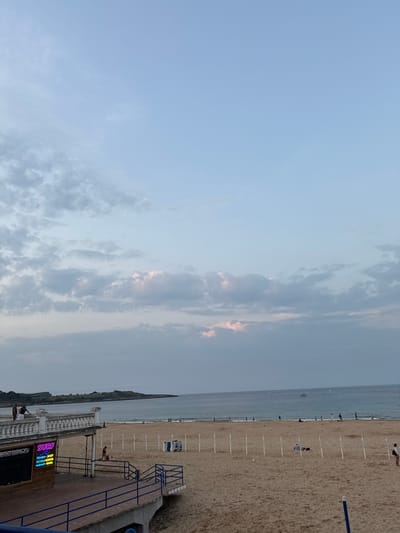 The image depicts a coastal scene at dusk, specifically the Primera Playa del Sardinero in Santander, Spain. The upper two-thirds of the vertical frame consist of a pale blue sky with scattered grey and white clouds, some exhibiting faint pinkish hues along the horizon, indicating low sun exposure.
The lower third of the image features a wide, sandy beach (Playa del Sardinero) extending from the foreground into the midground. The sea, the Cantabrian Sea, is visible beyond the beach, appearing calm with a gentle swell. Several small boats are discernible on the water in the distance. A green, rocky headland juts into the sea on the far left.
In the left foreground, an elevated concrete promenade with a white balustrade extends horizontally, supporting two indistinct figures. Below this promenade, a dark-fronted commercial structure is visible, featuring a blue and white neon sign that reads "SESSION SURF" with smaller text indicating "TIENDA - SHOP," "ESCUELA - SCHOOL," and "ALQUILER - RENTAL." A dark blue metal railing delineates access from the promenade to the beach level.
On the sandy beach, approximately a dozen individuals or small groups are scattered. One person is visible on the lower platform near the surf school sign. A series of white vertical poles are regularly spaced across a section of the beach in the midground. Near these poles, two folded blue and white beach chairs are stacked, and a small, light-colored conical object rests on the sand. Figures are also visible in the distance near the waterline. No specific interactions or detailed actions are clearly discernible for most individuals due to distance.