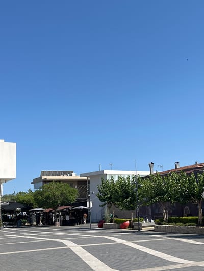 This is a wide-angle, eye-level, outdoor photograph taken on a bright, sunny day in the city of Limassol, Cyprus. The sky is a clear, vibrant blue with a single bird visible in the distance. In the foreground, a wide, paved plaza with a geometric pattern of dark gray and white tiles stretches across the scene.
Beyond the plaza, a row of buildings and trees forms the middle ground. On the left, a modern white building with a flat roof is partially visible. Adjacent to it, a multi-story building features a glass facade and a flat roof with solar panels. Further to the right, a two-story building with a terracotta tiled roof and chimneys stands. In front of these buildings, a line of trees with lush green foliage provides shade over a series of market stalls or shops. Several umbrellas, likely for the stalls, are visible. People are present, some browsing the stalls and others walking. Red, heart-shaped sculptures are placed on the ground near the trees and the plaza. A small water fountain is also visible.
The overall impression is of a lively, modern urban area with commercial activity and recreational spaces. The time of day is likely midday, given the bright sunlight and the clear sky. The environment appears clean and well-maintained.